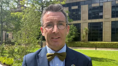 Minister Muir has short dark hair, round metal glasses and is wearing a navy suit jacket, blue shirt and green tie.