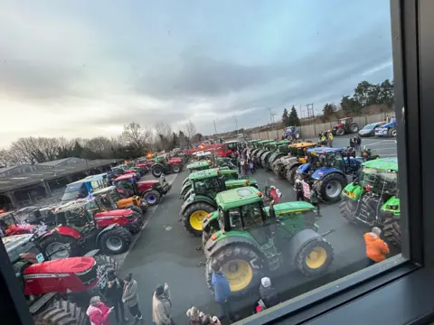 A view through a window down onto a car park which is filled with lines of tractors in various colours including red, green, and blue. The sky is grey and trees can be seen in the distance.