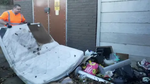 A man in an illuminous orange hi-vis top lifting a mattress which is among the illegal rubbish which has been dumped in Sefton. 
