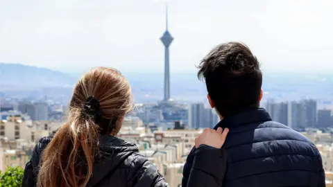 A woman and a man in black jackets look out over Tehran's skyline with a tall tower in the distance. They have their backs to the camera, she has long brown hair tied in a pony tail and her right hand rests on his left shoulder. 