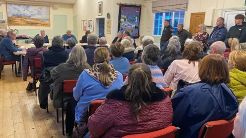 BBC A village hall is full of people seated in rows with more standing around the edge. At the front seven people are seated around a table 