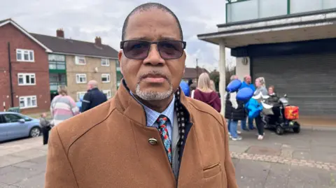 Kate Bradbrook/BBC Bishop Donnovan Allen standing outside a precinct looking at the camera. He wears lightly tinted sunglasses, a brown wool coat, a blue checked shirt and a patterned tie. He has a pale grey goatee beard and cropped black hair. Behind him is a group of people holding blue helium balloons.