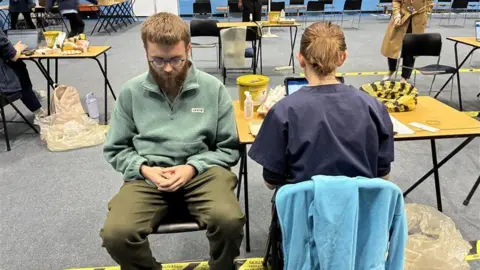 A student with glasses and a beard waits in a chair for a second jab.
