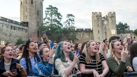 A young crowd stands at a concert with Warwick's historical castle as a backdrop. 
