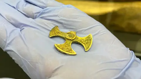 Leeds Museums and Galleries A small gold cross sitting on the palm of hand with blue gloves on