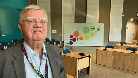 Amy Holmes/BBC A picture of councillor Steve Owen who is stood in the council chambers at Central Bedfordshire Council. He has a number of desks and chairs behind him, and is wearing a light blue shirt, a dark grey jacket and has glasses on.