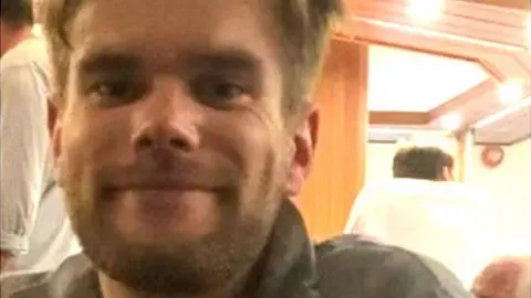 David Haw smiles at the camera in a small room, possibly a boat cabin. He has brown hair, cut short at the sides, and a beard.