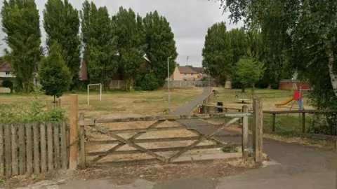 The play park in St Oswalds Road. There is a football pitch to the right and a enclosed play area with play equipment to the right. A large gate is seen on entry to the park, with a paved path to the right of it. There are residential buildings in the horizon.