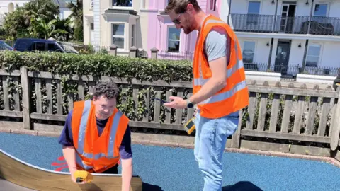 One man in an orange high vis vest kneels over a metal slide with a thermometer, while another man in a high vis vest and jeans stands over helping him.