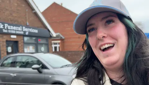 A woman with black hair, tinged with green highlights, smiles as she stands in front of a car park with commercial buildings in the background. She has a silver nose ring and is wearing a grey baseball cap and white coat.