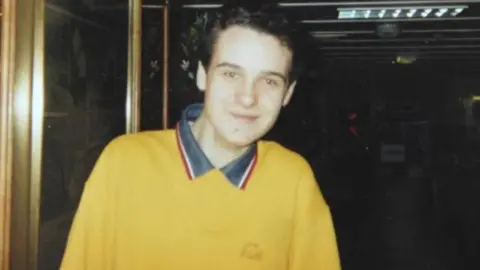 PA Media Alan Radford with black, curly hair, smiles at the camera wearing a yellow sweat shirt with a grey, red and white collar.