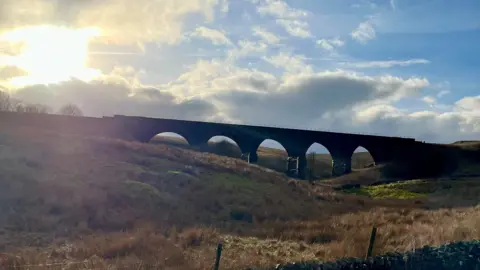 Weather Watchers/Sarah A skyline near sunset with a large viaduct silhouetted against the sky in Garsdale, Cumbria. The sky is blue with grey clouds and the sun is shining through. In the foreground there is a field and dry stone wall.