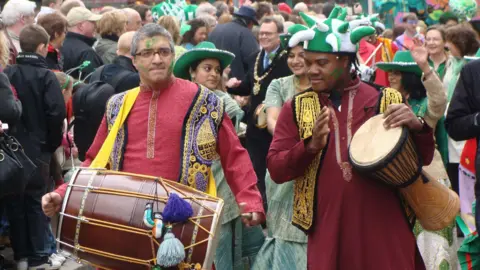 A colourful street parade featuring two individuals in the foreground playing traditional drums. One person holds a large dhol drum and wears a red kurta with a decorative vest, while the other plays a smaller hand drum and is dressed in a maroon kurta with a similar vest and a green-and-white jester-style hat. The vibrant setting shows a lively crowd in the background, many adorned in green accessories, suggesting a festive celebration.