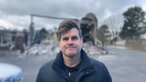 Louis Ludik, who has short dark hair parted to the side, stands in front of a building destroyed by fire