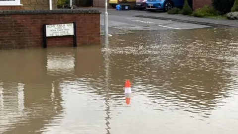 A view of a flooded suburban street. A street sign reads Home Farm Way. There is a cone sticking out from the middle of the water.