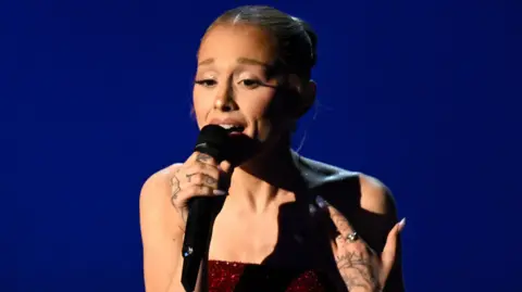 Getty Images Ariana Grande in a red sparkly dress singing into a handheld microphone at the Oscars in March 2025