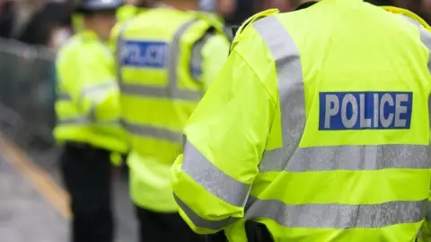Three police officers standing outside wearing yellow fluorescent jackets with silver stripes and silver writing in a blue box which says POLICE.