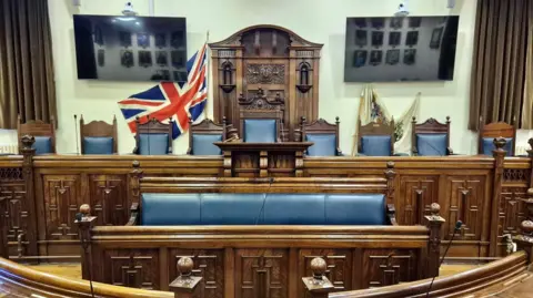 Rochford District Council A council chamber, showing a Union Flag at the back, wooden seats and benches, with chairs with blue leather on then, and a bench, with blue leather on. There are two television screens to the left and right.