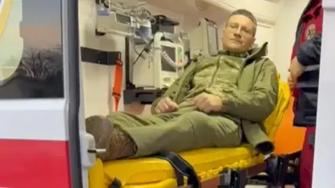 Maryna A man dressed in army khaki green uniform, lies on an yellow stretcher inside an ambulance. The man is sitting up and is wearing glasses.