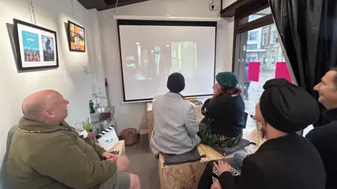 The inside of the Creative Place, a small rectangular building with the market square visible through curtains around a window. Several people are sitting inside watching a screen on which is a man in a suit. There are picture frames on the walls.