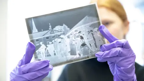 Inspire Culture A woman wearing latex gloves holds up a photographic negative between her face and the camera