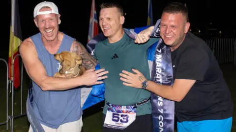 World Marathon Challenge Three men - including one holding a small dog - laughing in the dark after crossing the finish line of a marathon. 