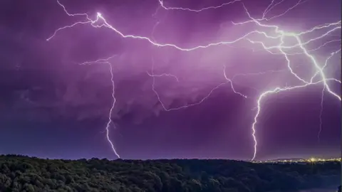 Matthew Harris Very bright white lightning strikes seen in a purplish sky with wooded trees beneath