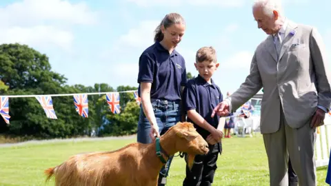 Andrew Matthews/PA The King is dressed in a grey suit. He is pointing to the golden goat as he talks to the young handlers who are dressed in blue tops and jeans