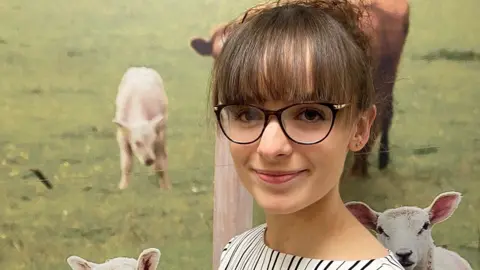 AHDB Photograph of Lloyd, who is looking into the camera and smiling slightly. She is pictured indoors, in front of a wall which has a large photo of sheep in a grassy field printed onto it. Lloyd is wearing a black and white striped top and a pair of glasses. 