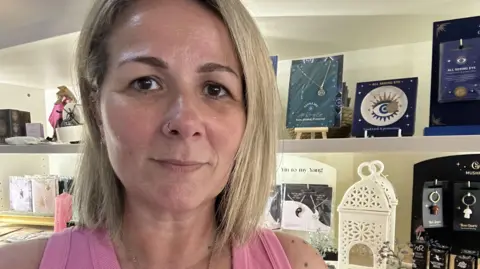Ben Parker/BBC A woman with blonde hair looking at the camera, she has a pink top on. Items are on shop shelves behind her.