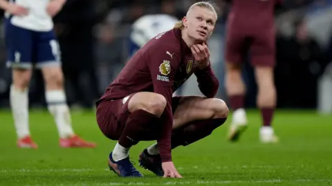 PA Media Manchester City striker Erling Haaland crouches down on the pitch at the Tottenham Hotspur Stadium during the Premier League match against Spurs. He is scratching his neck. He is wearing City's maroon-coloured away kit. Other players from both teams are walking around in the background.