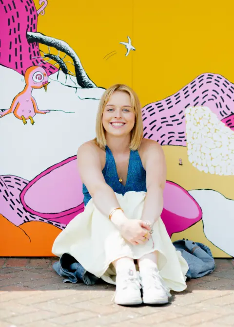 Blake Watson A woman with blonde hair sitting on the ground against a brightly painted wall smiling at the camera