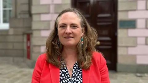 Deputy Hilary Jeune is standing in front of Jersey's States Assembly building. She is wearing a black and white shirt with a pink blazer and green earrings. She is looking into the camera.