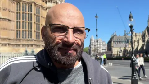 Khalid Hussain is bald with glasses and a full beard. He stands on a pavement at Westminster.