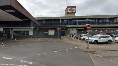 Google A wide grey and brown building with blue railings and a red national rail logo at the top. A number of cars and red no entry signs surround the building.
