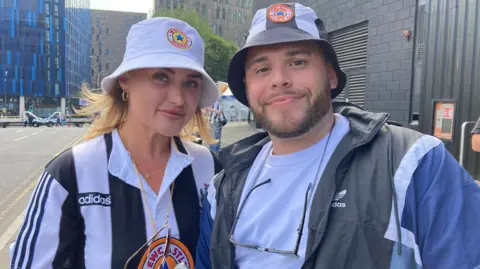 A woman and man close to St James' Park Newcastle before Sam Fender's gig. She has blonde hair and wears a Newcastle United home shirt. Both wear Newcastle themed hats and sunglasses on chains around their necks. The man has a trimmed black beard and moustache.
