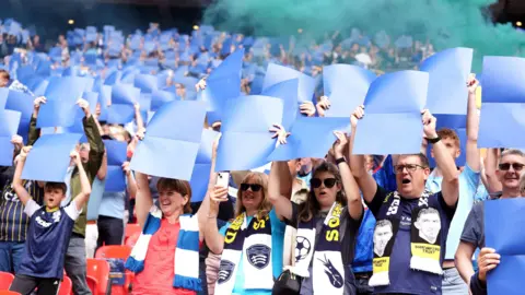 Adam Davy/PA Southend supporters stand up in their seats at Wembley stadium, holding large blue sheets of paper above their heads. Many wear their club's scarves and kit. There is blue smoke wafting above them.