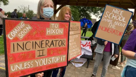 A group of people, one wearing a mask, are holding signs while protesting in a park, next to a park bench. Slogans are written on the signs, 