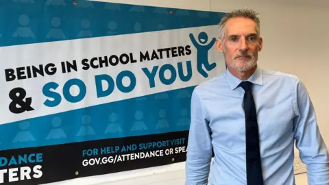 A middle-aged white man in a light blue shirt and dark tie stands beside a large blue attendance campaign banner on a white office wall. The banner reads, “Being in school matters & so do you” and includes the Attendance Matters logo and a link for attendance support information.