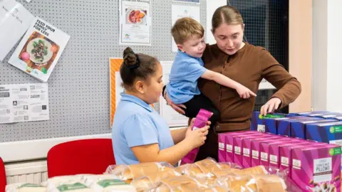 Matt Grayson / Save the Children A woman holding a young child with another child stood next her. In front is a large table covered in food items