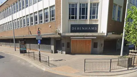 Google Google streetview image of the former Debenhams store, a large mid-20th century building with brown-brick walls and two long rows of windows separated by white panels. On the ground floor, metal shutters cover the windows. Wooden boards cover the double doorway.