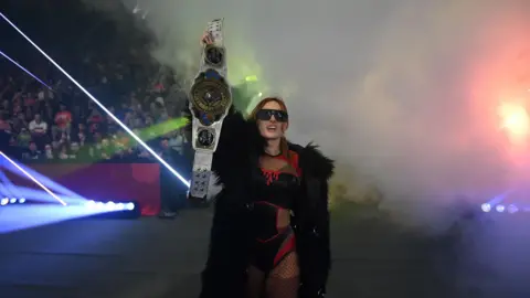 Getty Images A female wrestler is holding a championship belt. She has black glasses and a long black coat on with her wrestling costume. There is smoke t the right side of her. 