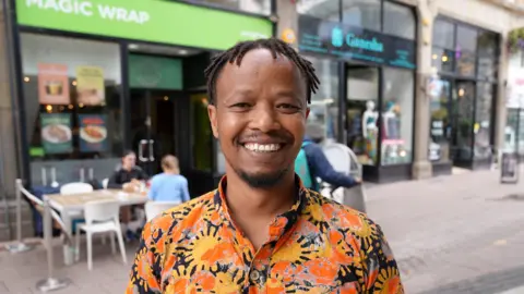 Fadhili Maghiya smiling at the camera on Cardiff Queen Street. He is wearing an orange, yellow and black patterned shirt