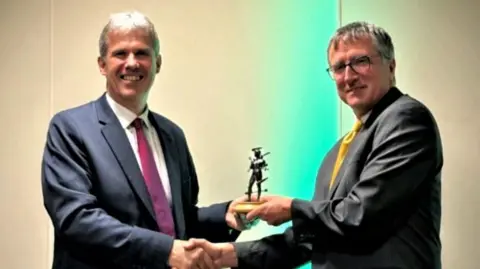 CUH Dr Rod Mackenzie smiles at the camera as he is passed his award by Professor Tim Coats. Both men wear suits and have short grey hair. They each hold the award in one hand as it is passed over. 