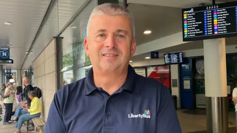 Kevin is wearing a navy blue LibertyBus polo shirt stands outside a bus station. Behind him are passengers waiting near benches, covered walkways with platform signs, and digital departure boards displaying bus routes and times. The station entrance and bus bays are visible in the background.