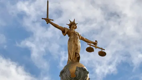 A golden statue of Lady Justice, above the Old Bailey (not in shot) stands tall on an ornate pedestal, holding a sword in one hand and scales in the other. She is set against a cloudy blue sky.