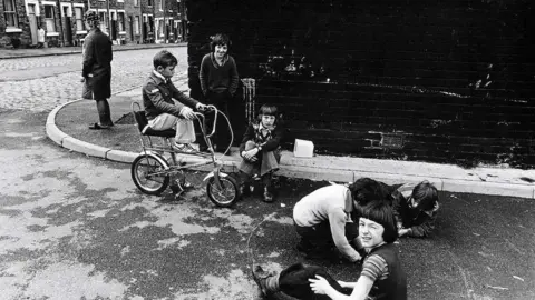 A black and white photo of six children playing on a street coroner while a woman with rollers in her hair stands on the pavement. 