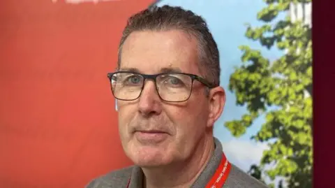 Bernard Trainor with brown/grey short hair wearing black framed glasses standing in front of a NIPSA poster. He is also wearing a grey t-shirt and a NIPSA red lanyard.