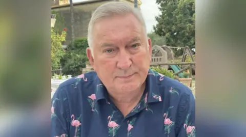 A head and shoulder image of a man - Dorian Jones - with grey hair wearing a navy blue skirt with pink flamingo on it looking down the camera lens with a white trees and a park in the background.