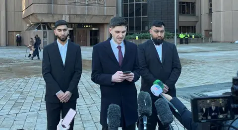 Mohammed Fahir Amaaz and Muhammed Amaad stand outside Liverpool Magistrates' Court wearing black suits either side of their solicitor who is reading a statement to the media from his phone. 